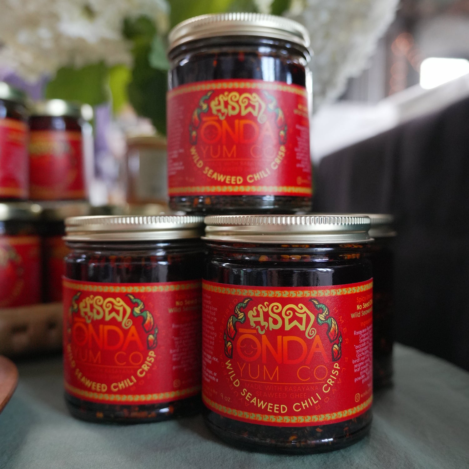 Three glass jars of Onda Yum Co. Wild Seaweed Chili Crisp arranged on a table, with a blurred background of white flowers and a green tablecloth.