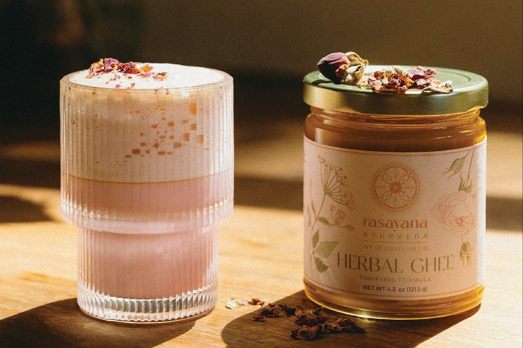 Glass of layered drink with a jar of rasavana herbal ghee on a wooden surface.