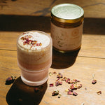 Glass of layered drink with foam and dried herbs next to a jar of herbal ghee on a wooden surface.