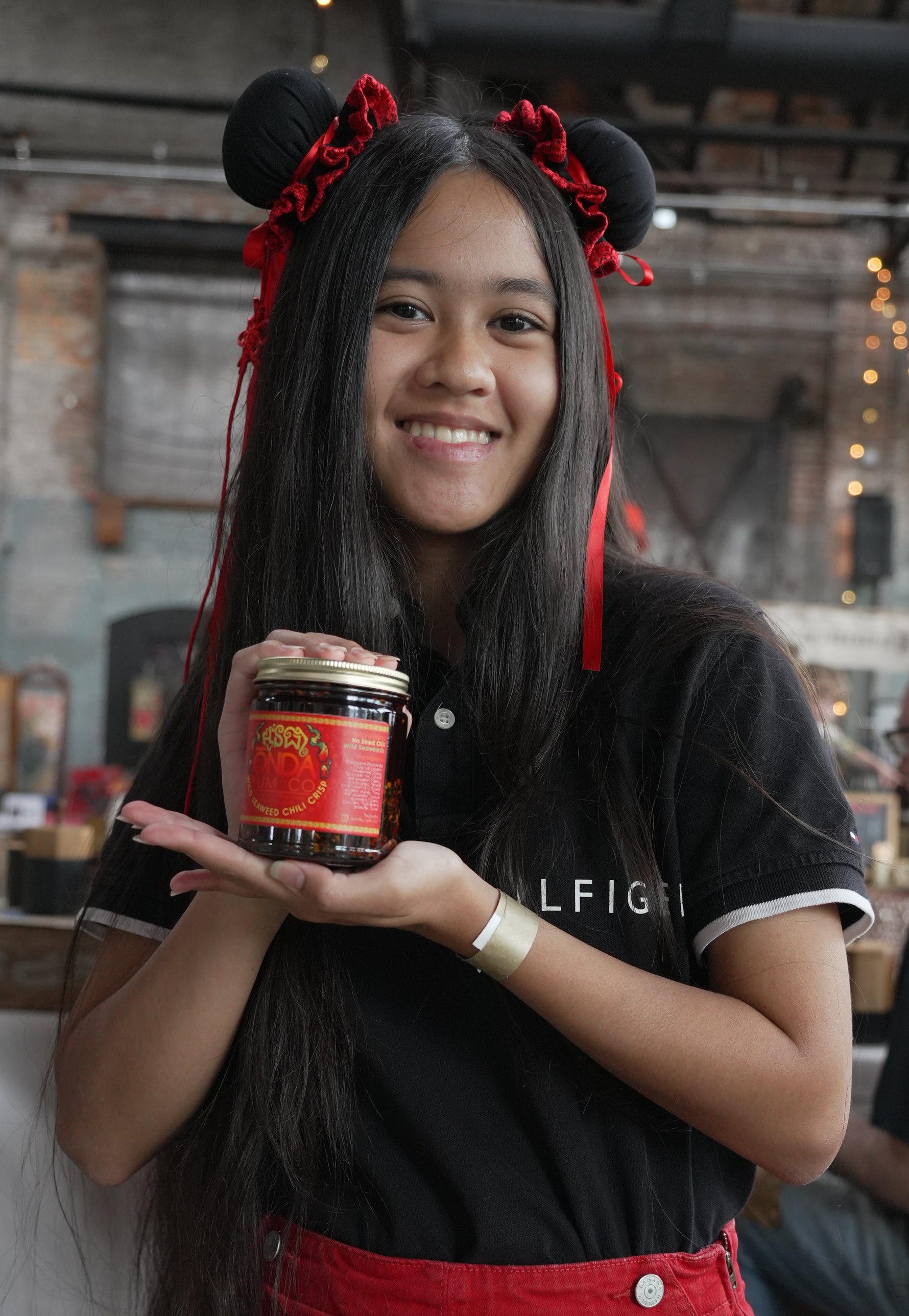 A smiling woman with long black hair and red headbands holds up a jar of ONDA Yum Co. Wild Seaweed Chili Crisp at  a market. She is wearing a black T-shirt with white trim and a red apron.