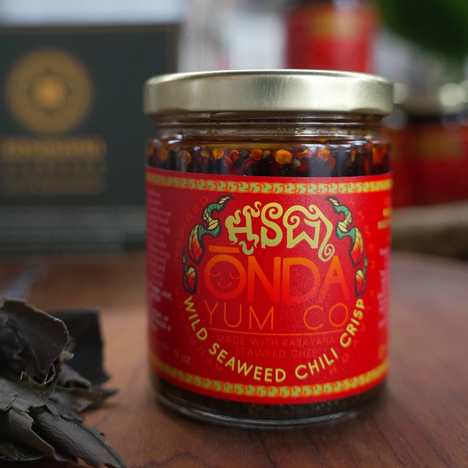 A glass jar of ONDA Yum Co. Wild Seaweed Chili Crisp with a bright red label and gold lid, set on a wooden surface with a blurred background.